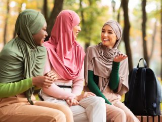 Three Muslim Arab Women In Headscarf Talking Sitting On Bench Outside In Park Enjoying Autumn Weekend. Female Friendship, Happy Islamic Students Enjoying Conversation Outdoors Concept